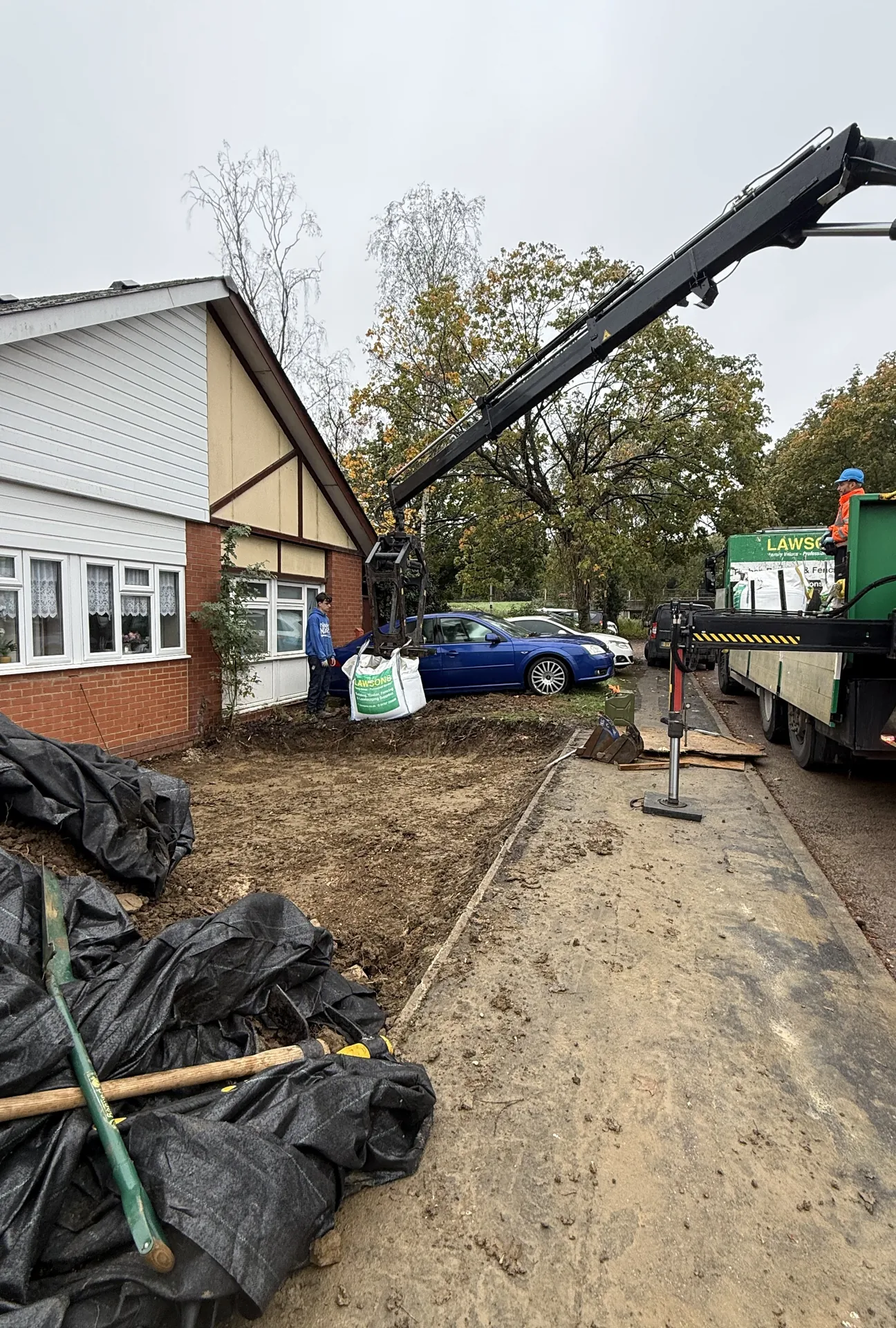 A crane lorry unloading bags beside the drive, with black sheeting heaped on one side and the stone base on the other.