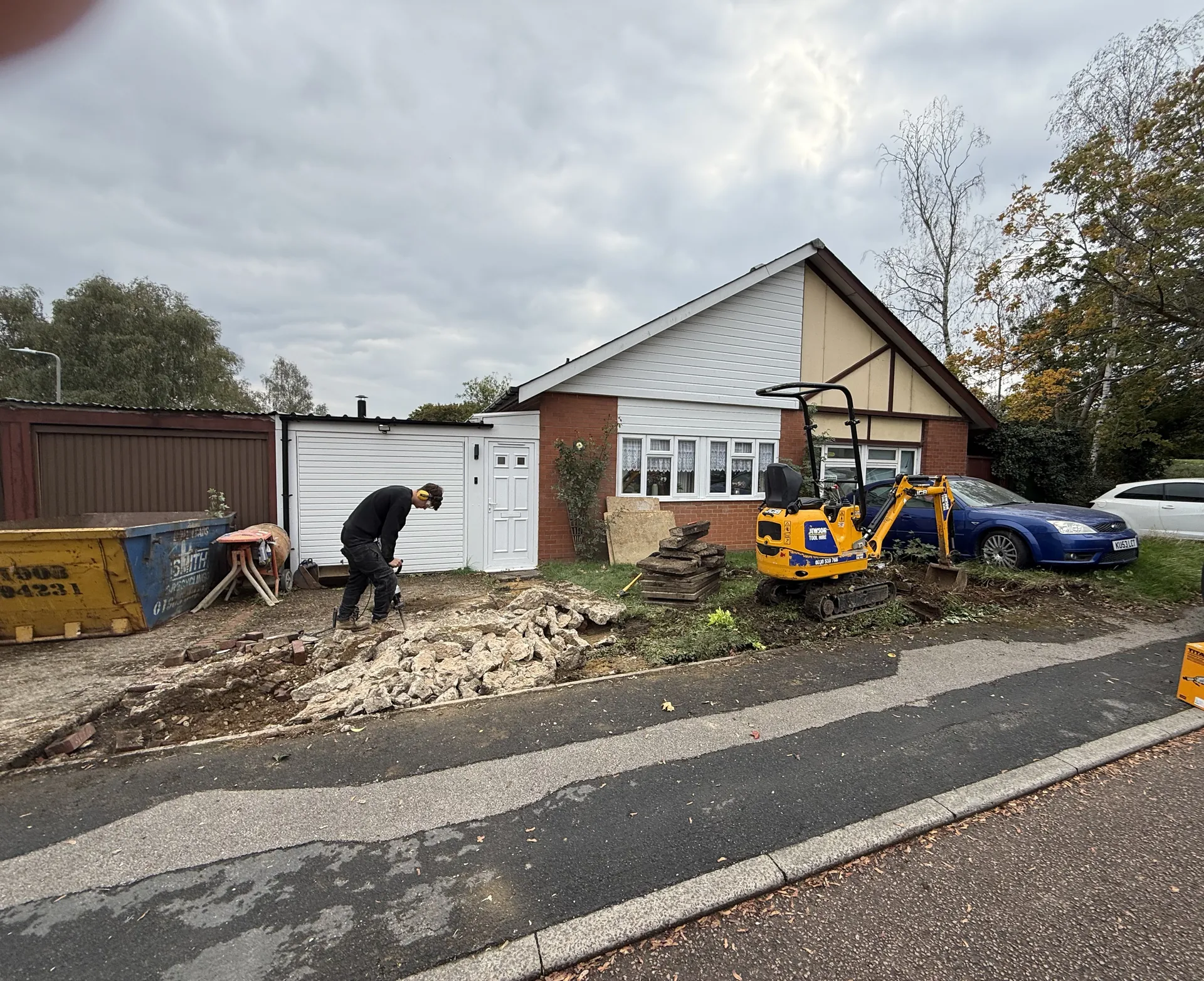 A small digger working at the front of the bungalow, with broken-out concrete piled by the road.