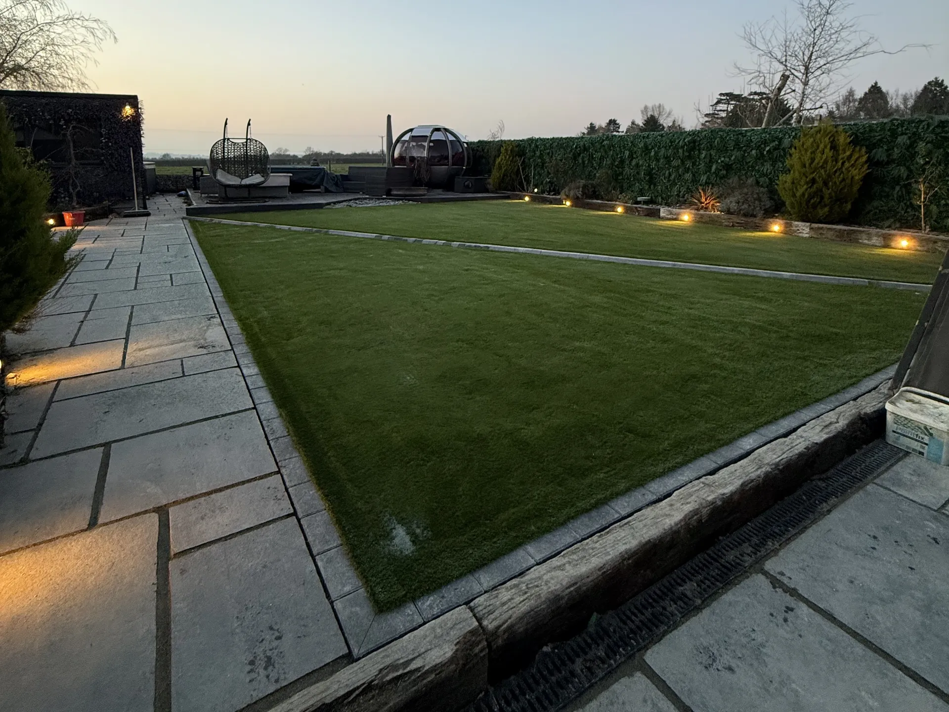 The relaid lawn from the same spot at dusk, with charcoal edging along the paving and lights along the far border.