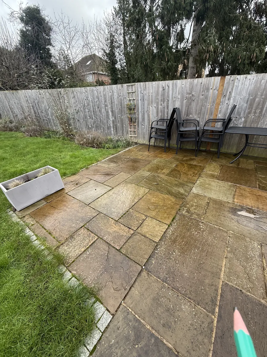 The stone patio in February, with moss and weeds in the joints and a granite sett edge along the lawn half hidden by grass.