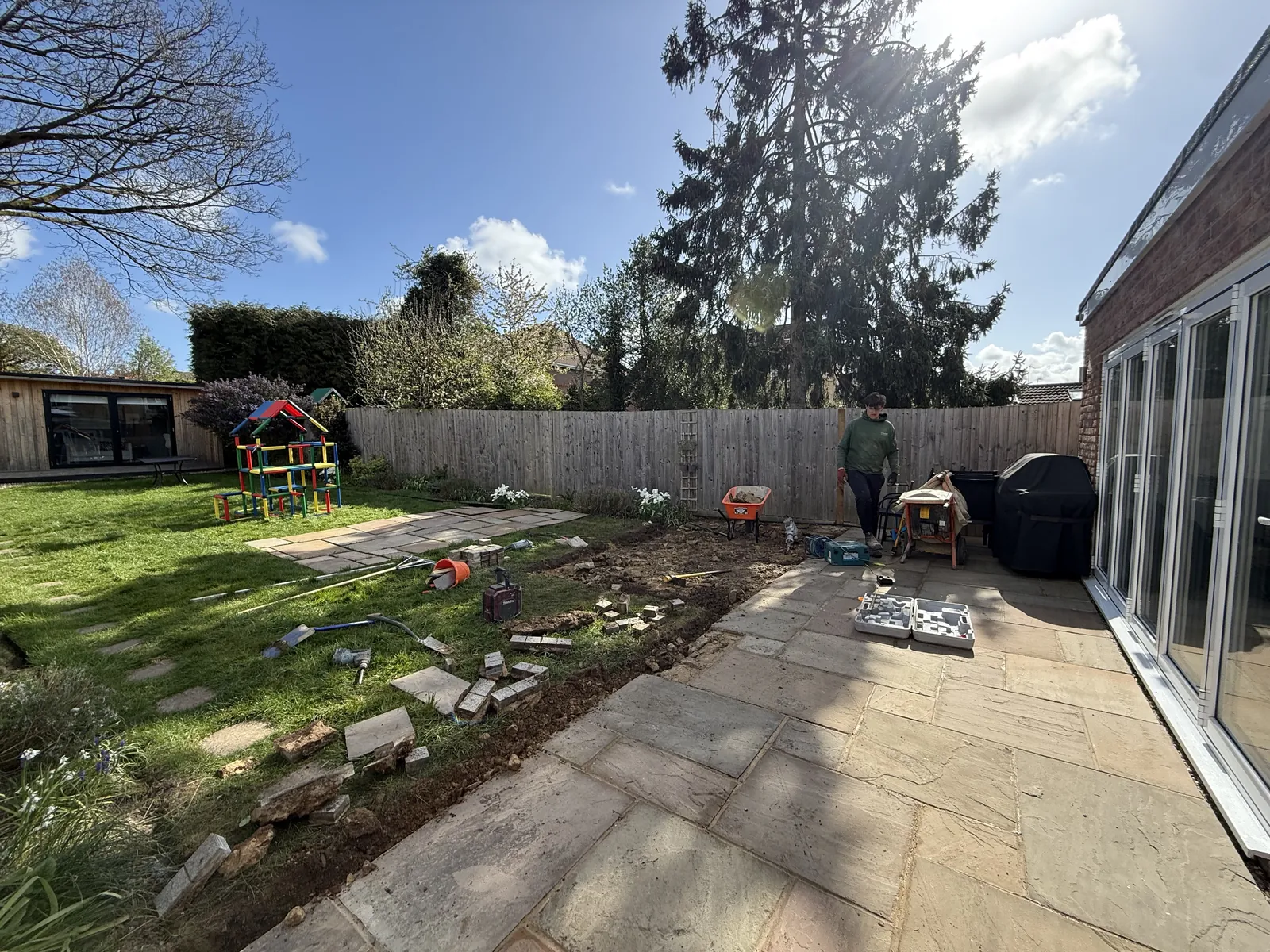 Mid-job, a strip of the patio lifted along the lawn, with old slabs laid out on the grass and the granite setts and rubble beside them.