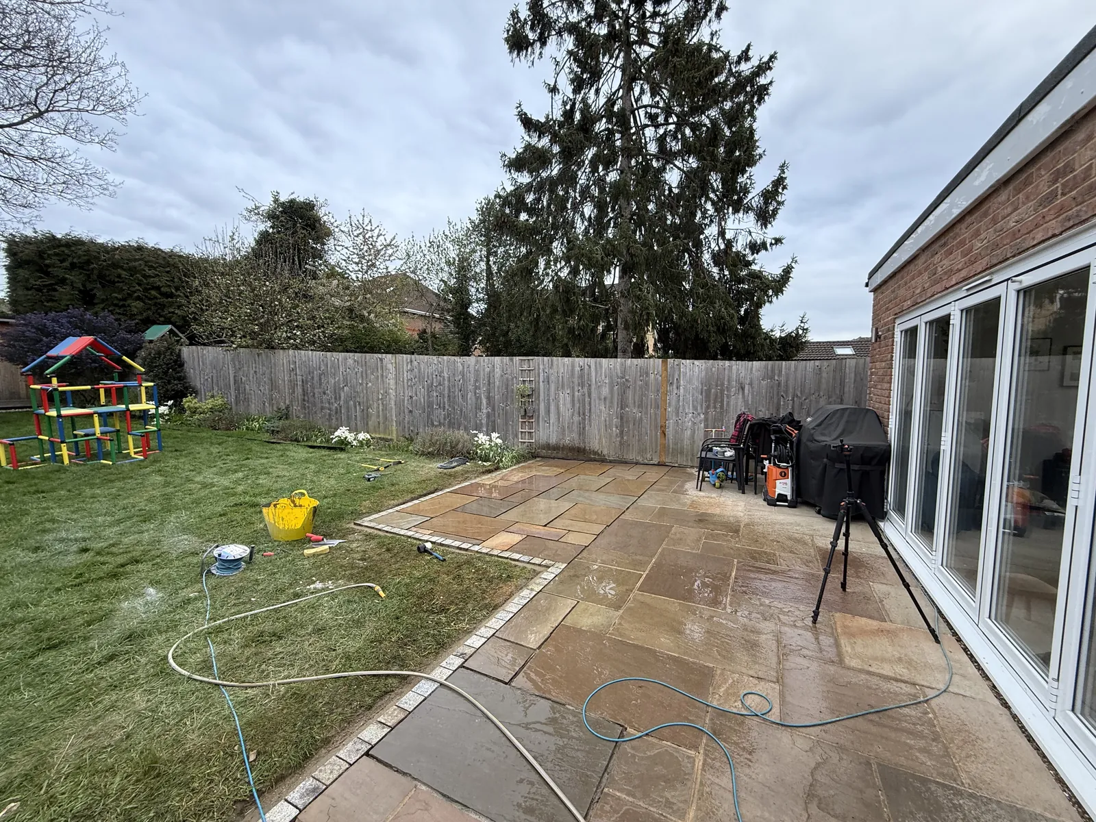 The finished stone patio along the back of the house by the bifold doors, with the relaid section and granite edge beside the lawn, and hoses still out.