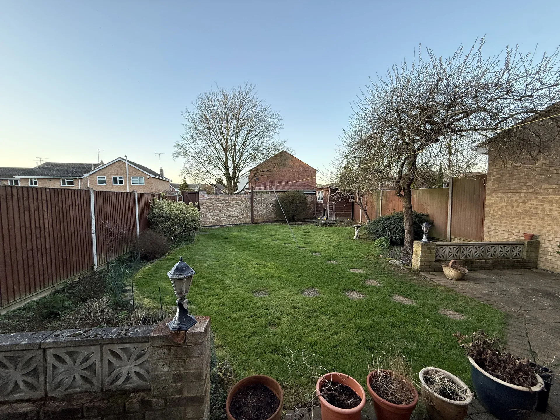 The old garden from the patio, with a screen-block wall and plant pots in front, stepping stones across the lawn and a bare tree.