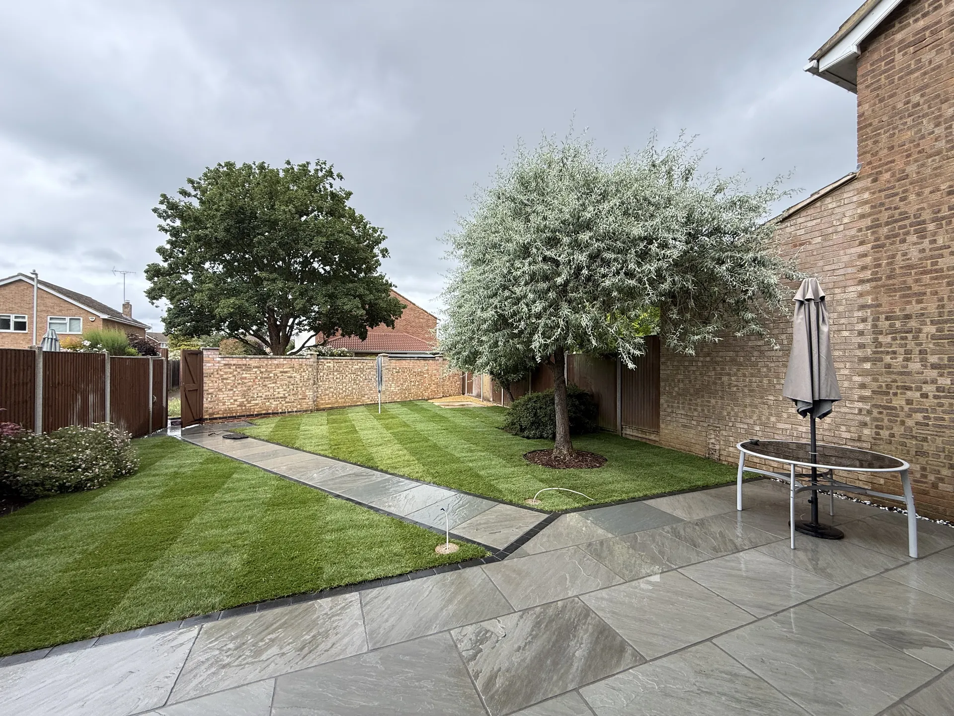 Grey riven sandstone patio with charcoal edging, and a sandstone path running past new lawn to the back wall.
