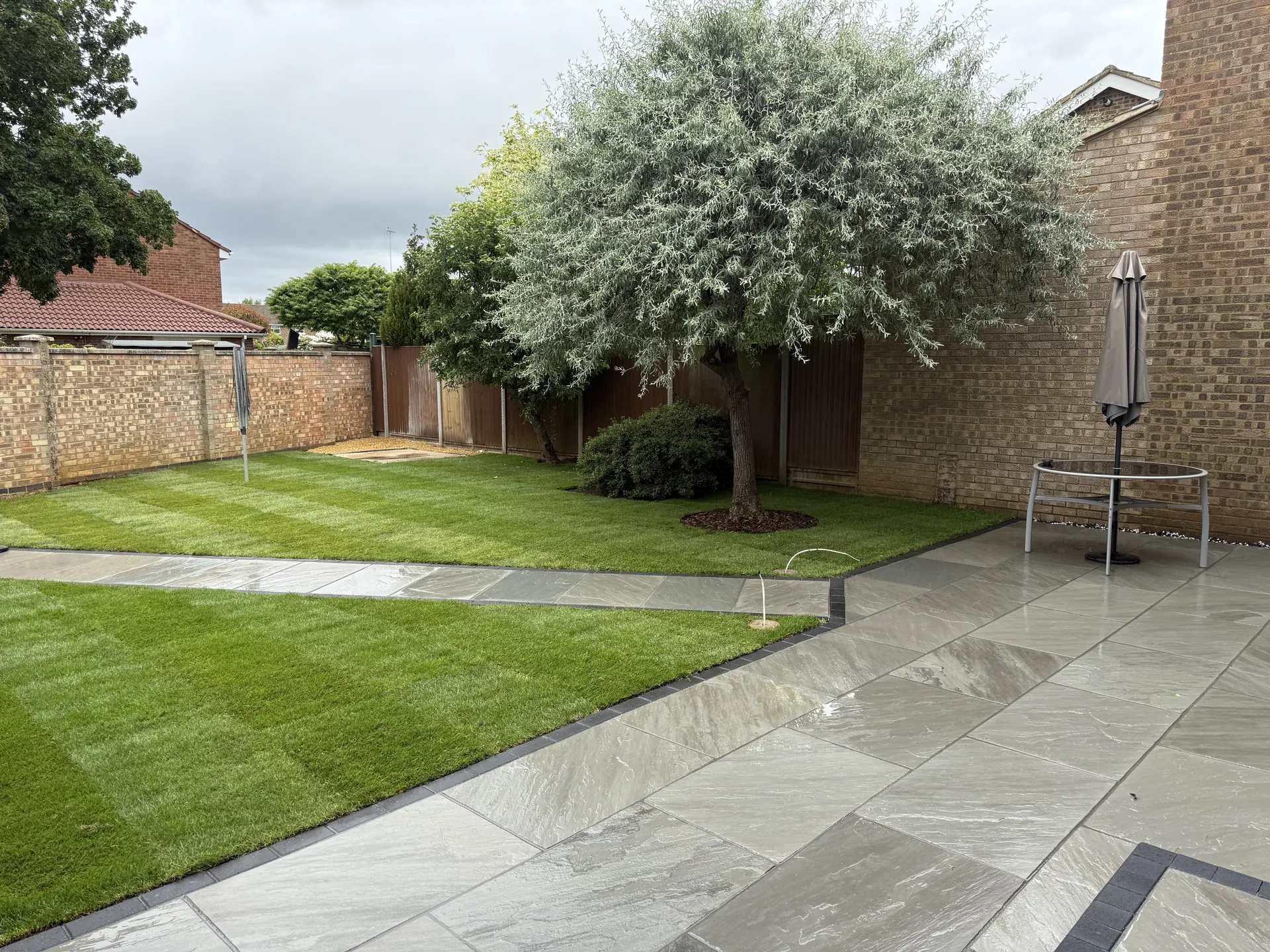The grey sandstone patio meeting the path, both edged in charcoal, with new lawn and a tree by the back wall.