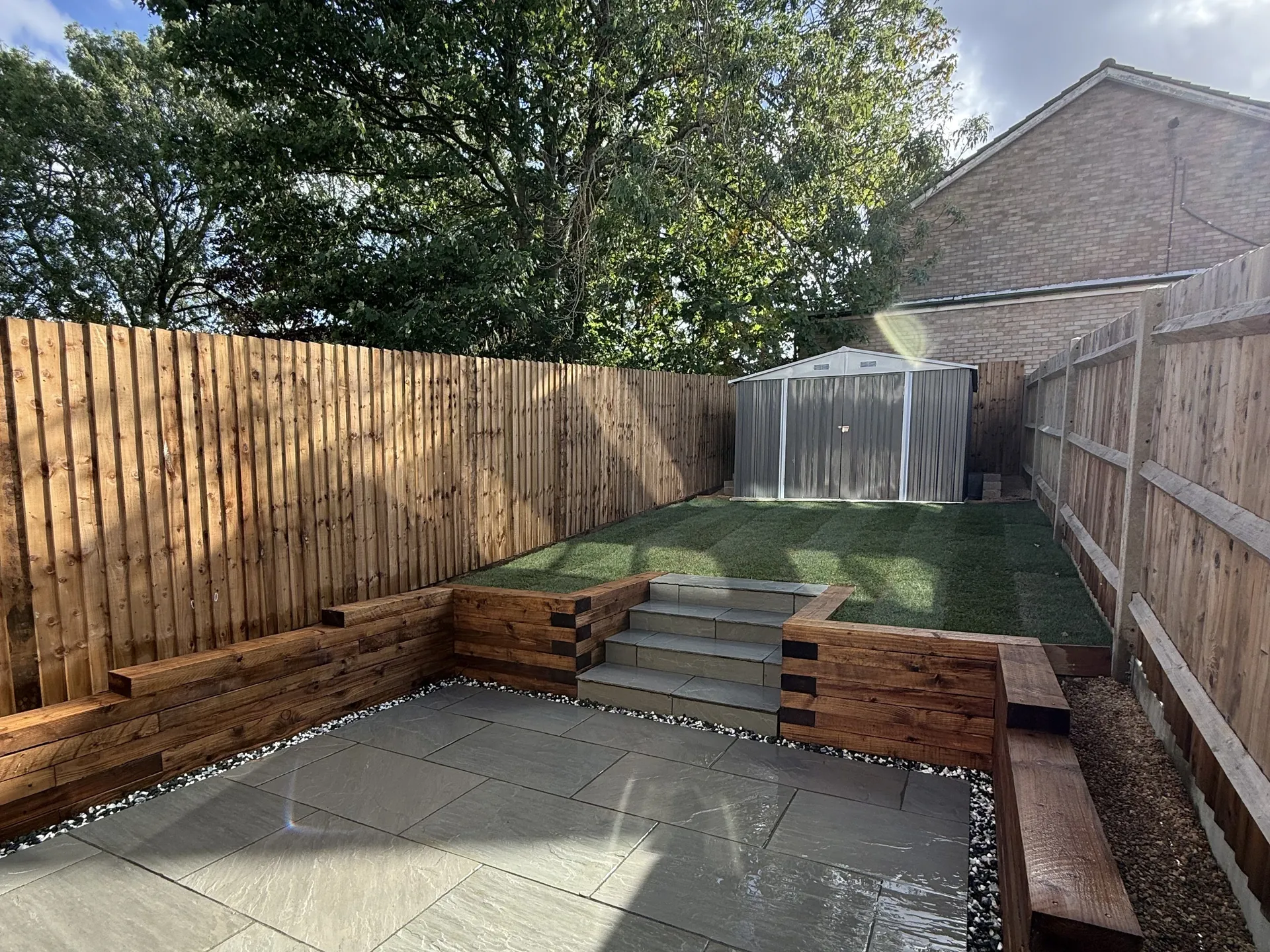 The finished garden from the corner by the house: a Kandla Grey sandstone patio, sleeper walls, sandstone steps up to a new lawn, and the shed at the top.