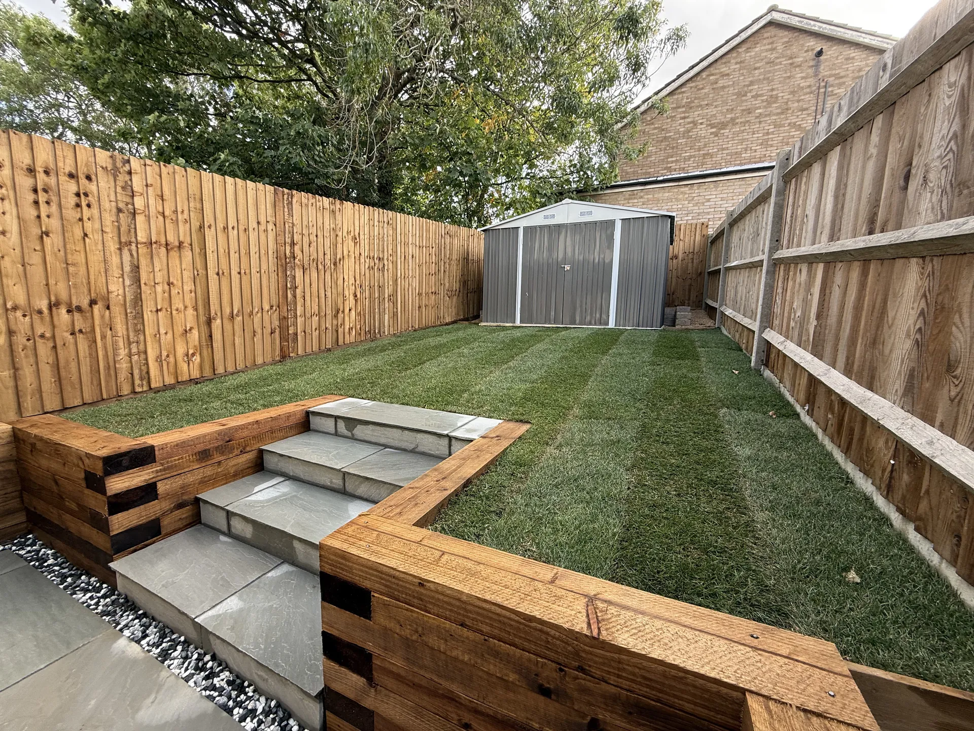 The finished garden from the patio corner, with sandstone steps between sleeper walls leading up to a striped new lawn and a grey metal shed.