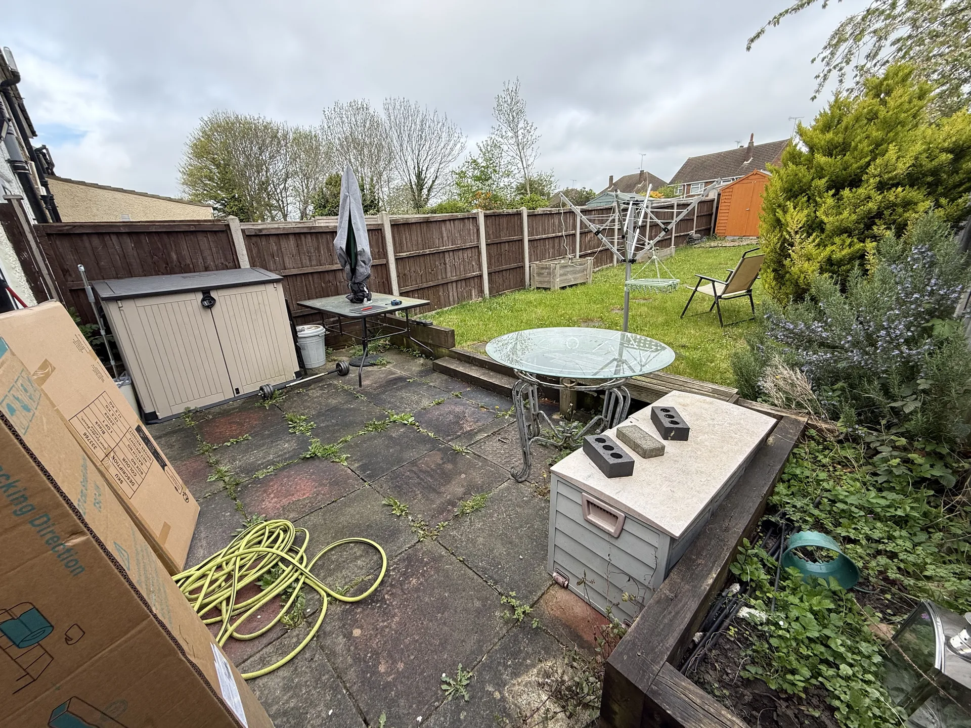 The old patio behind the house, with dark, stained slabs, weeds in the joints, old timber sleepers along the lawn and garden furniture and boxes on top.