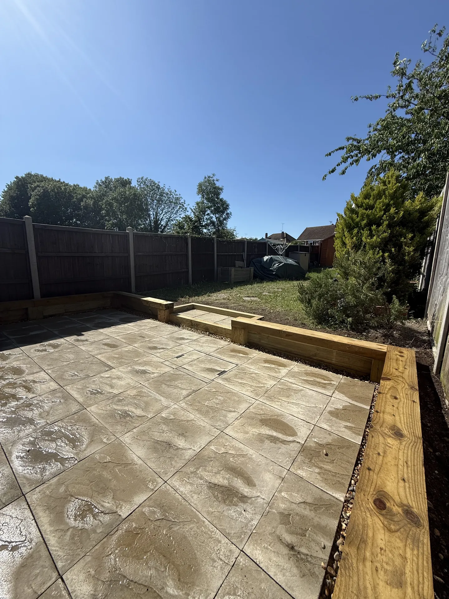 The finished beige patio seen from the house, with the new sleeper edging along the side and back and the lawn beyond.