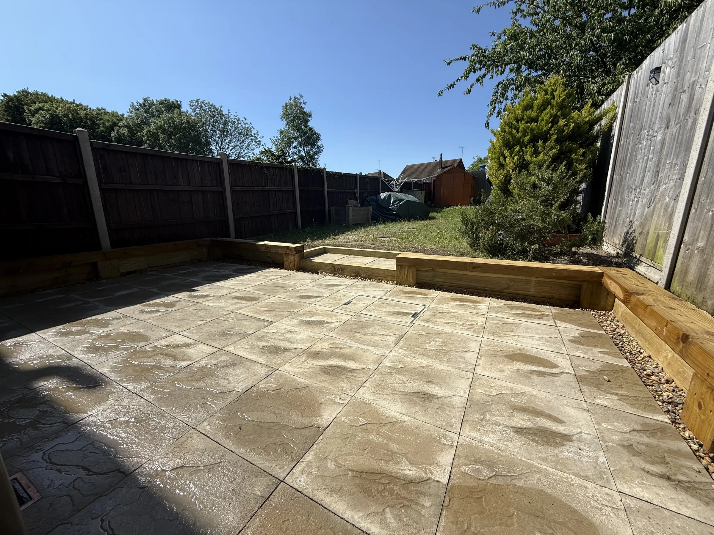 New beige concrete slab patio in a Luton garden, edged with new green timber sleepers, with a paved step up to the lawn.