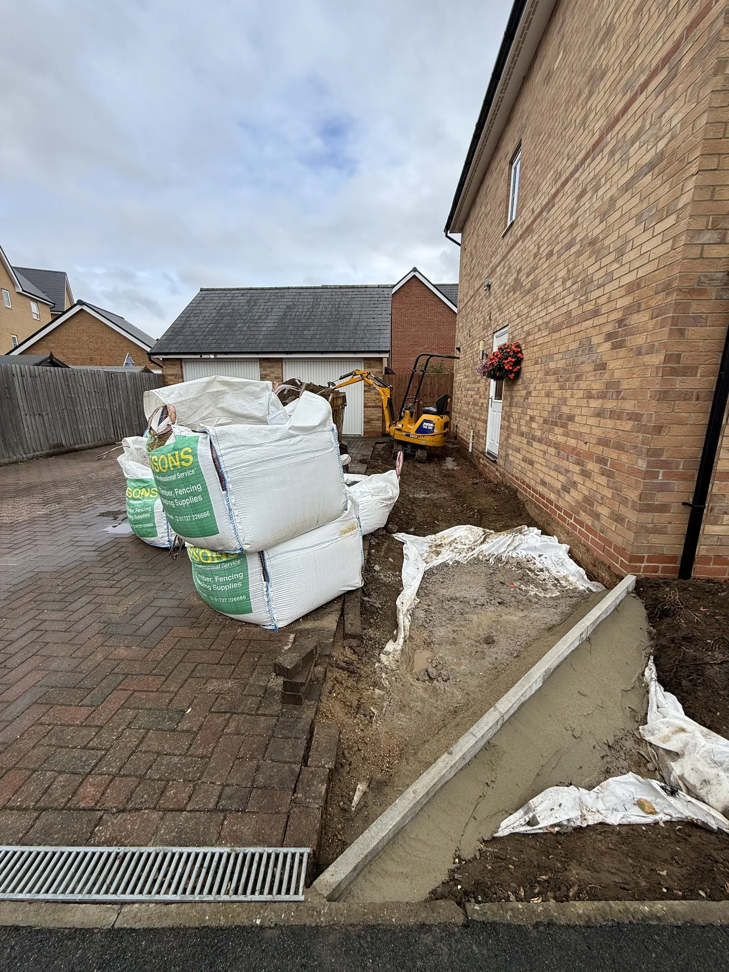 Mid-build beside the house, with the ground dug out, the new angled front edge set in mortar, bulk bags of materials on the old drive and a mini digger behind.