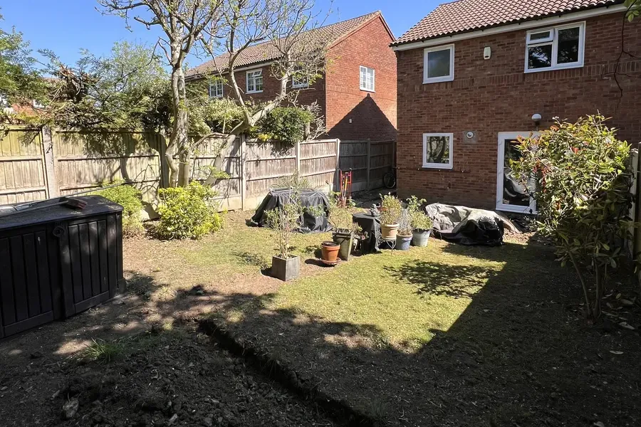 A Luton back garden left half dug by another firm, with bare churned soil in the foreground, a worn lawn, pots and covered furniture, and the brick house and back doors beyond.