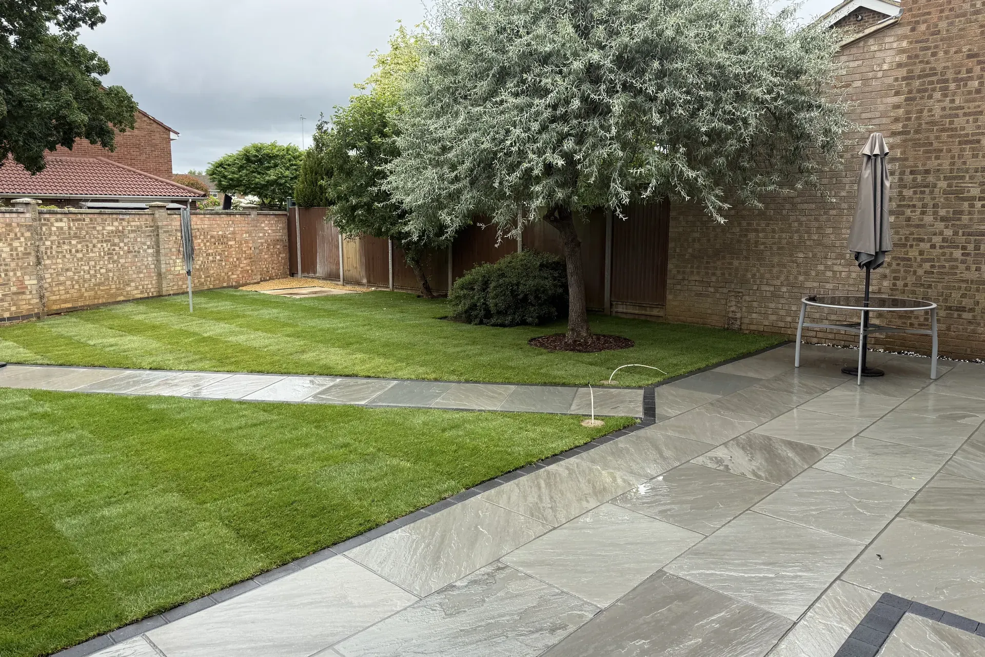 Our grey riven Indian sandstone patio in Leighton Buzzard, edged in charcoal, with a sandstone path, a new striped lawn and a tree against the back wall.