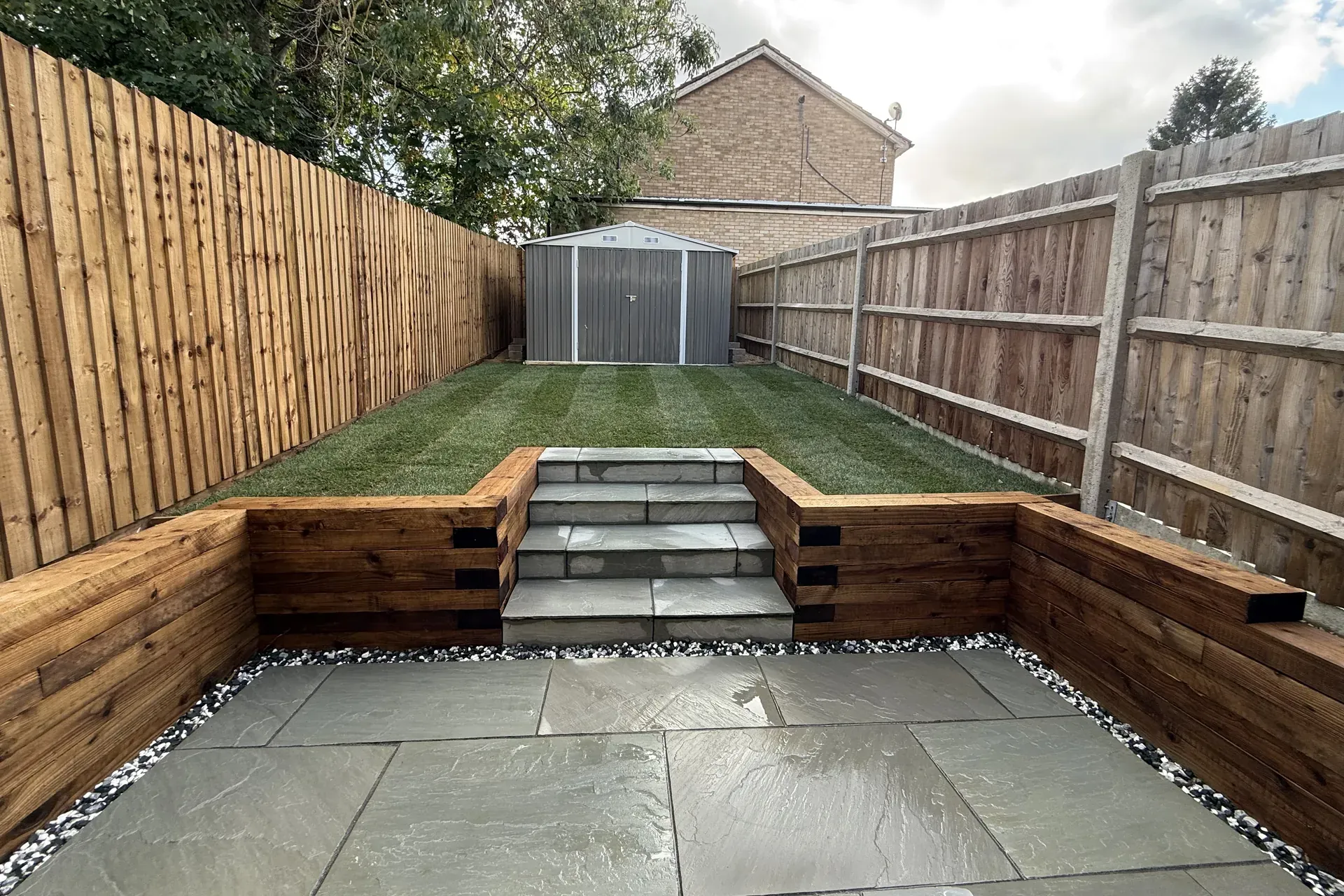 Our Luton garden from the back door, with a grey sandstone patio, timber sleeper retaining walls on three sides and sandstone steps up to a sloped new lawn and shed.